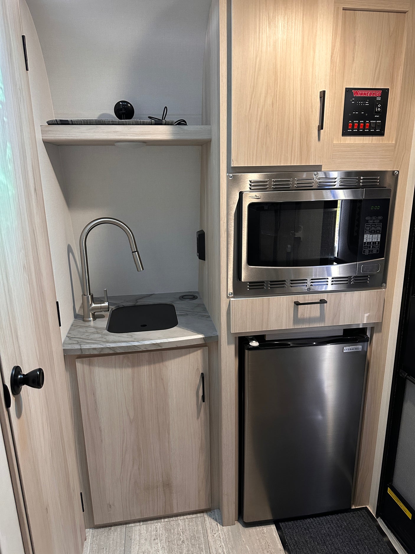 Compact RV rental kitchenette with a stainless steel microwave, mini-fridge, sink, and Winnebago control panel.