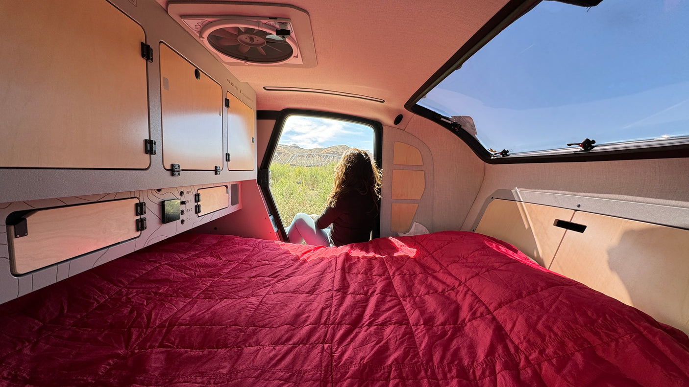 Cozy RV rental interior: A person enjoys mountain views from the open hatch, with a comfy bed and storage.