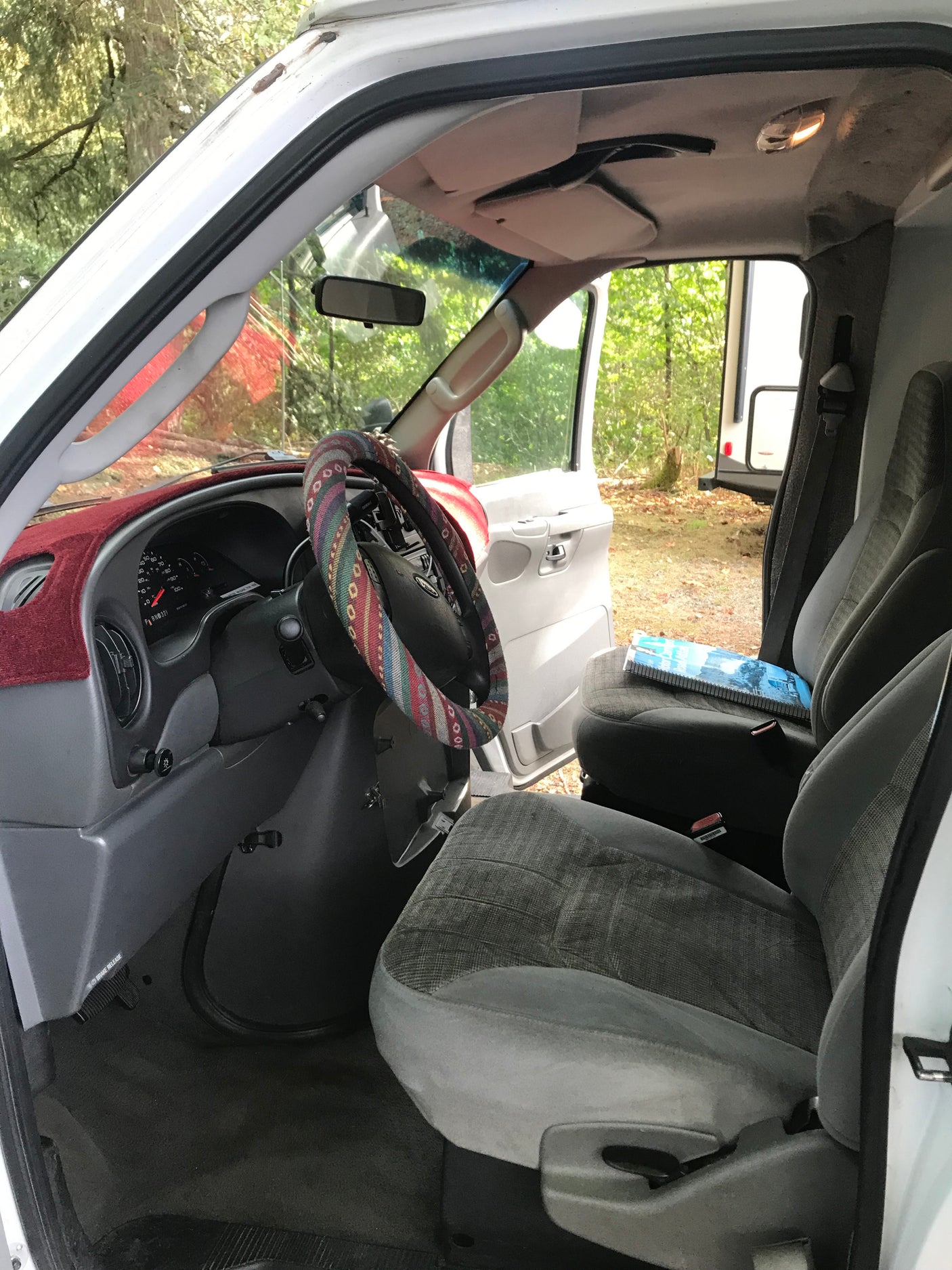 Interior of an RV rental: driver's seat, dashboard with a colorful steering wheel cover. Trees and another camper are visible outside.