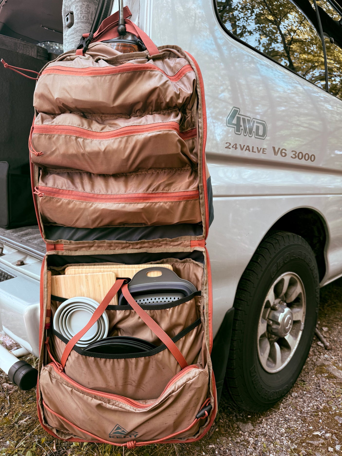Kelty camping kitchen organizer with bowls, cutting boards, & colander, hanging on a white 4WD RV rental van.