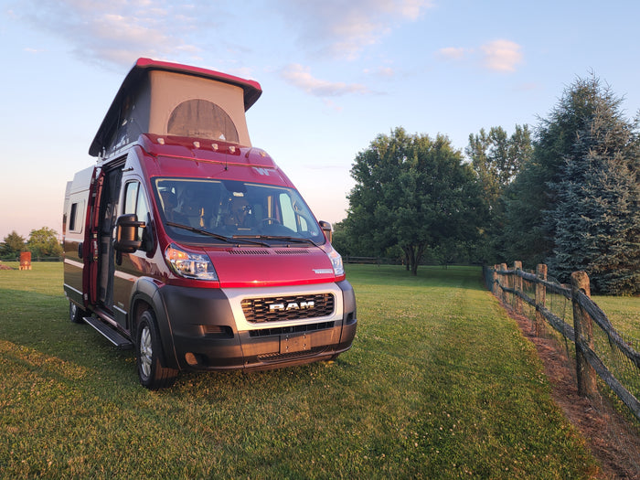 Red Winnebago camper van RV rental, pop-top open, ready for adventure on a grassy field at golden hour.