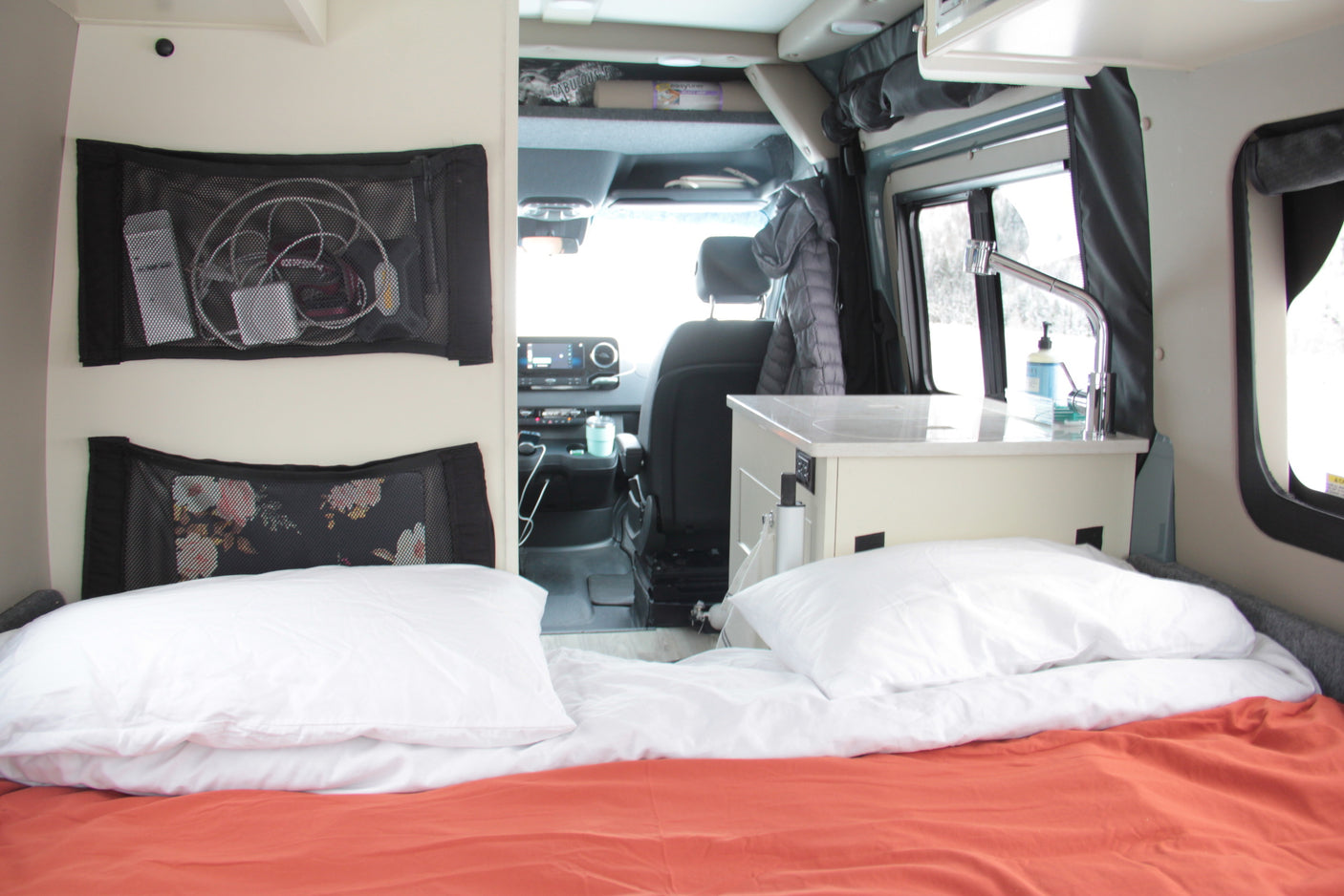 Explore this RV rental's interior: cozy bed, mesh storage with electronics, and a small kitchen counter with sink. Driver's cabin visible.