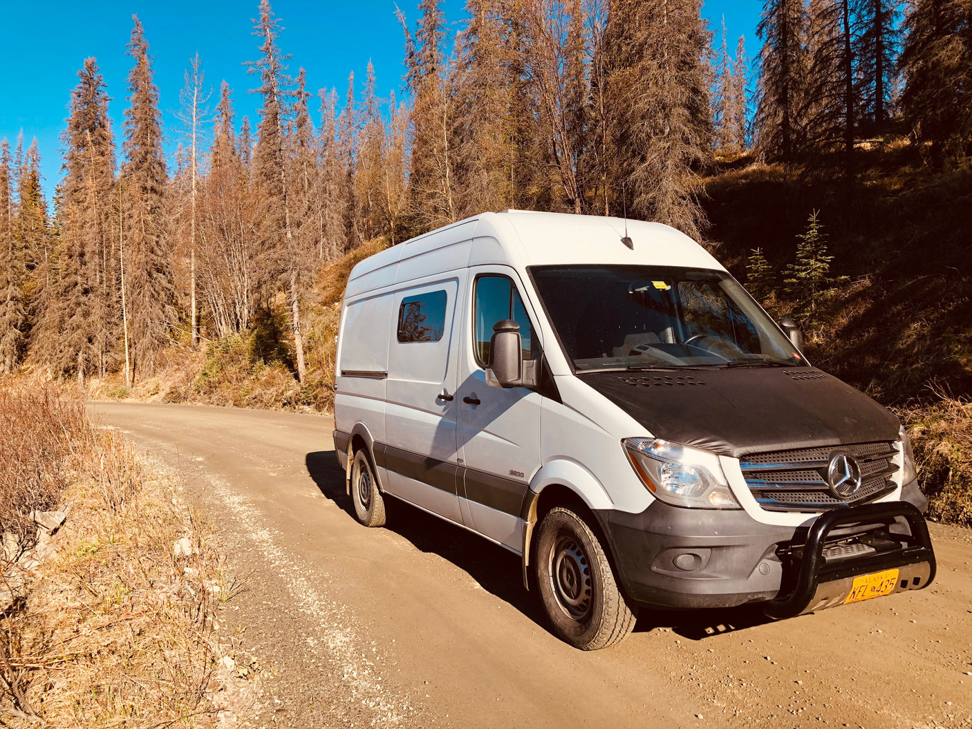 A white Mercedes Sprinter RV rental van with a bull bar, on a dirt road amidst dry trees under a blue sky.