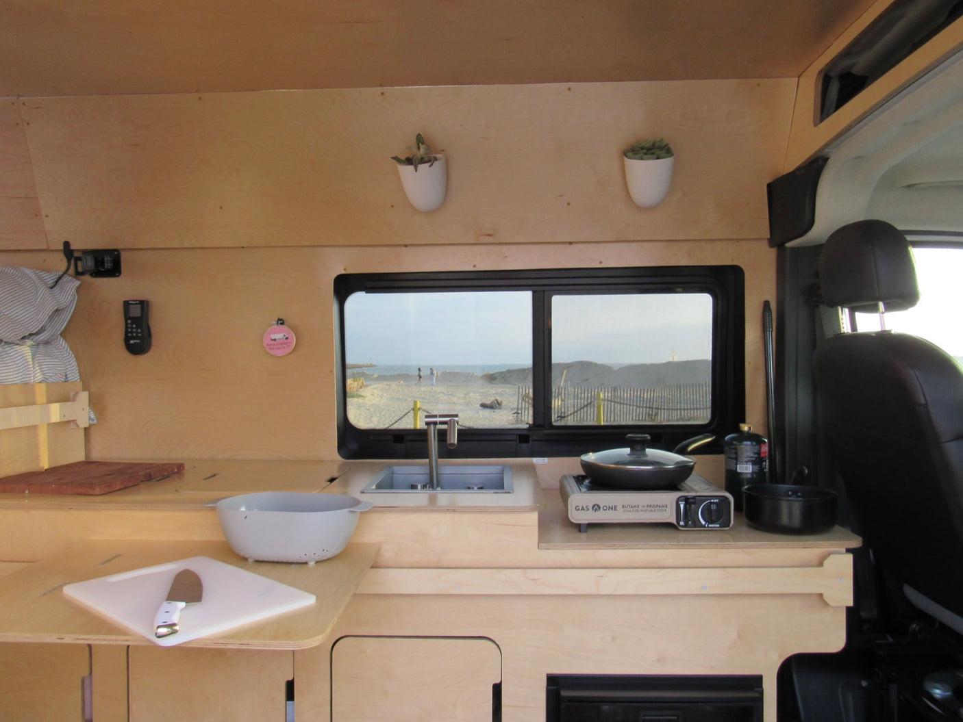 This RV rental's compact kitchen boasts a sink, portable stove, and a beautiful window view of a sandy beach.