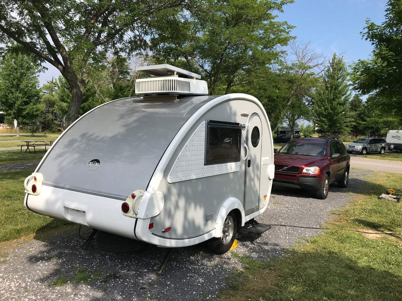 A sleek gray T@B travel trailer, an ideal rv rental, set up on a sunny campground site with a red SUV tow vehicle.