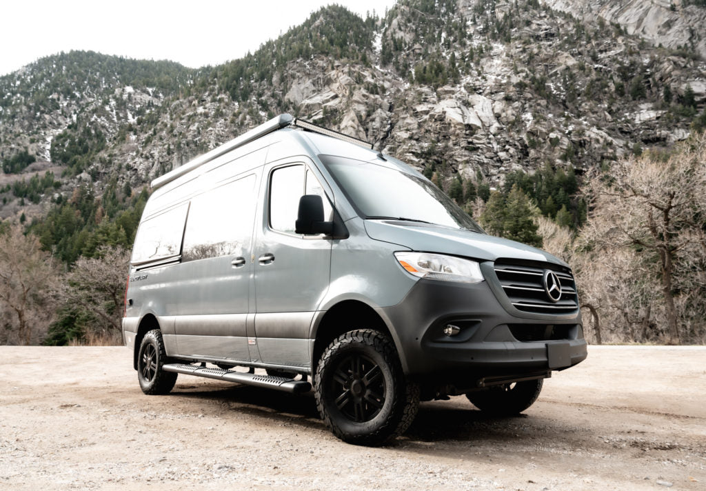 Gray Mercedes Sprinter adventure van, equipped for off-road RV rental mountain trips, parked on a dirt trail.