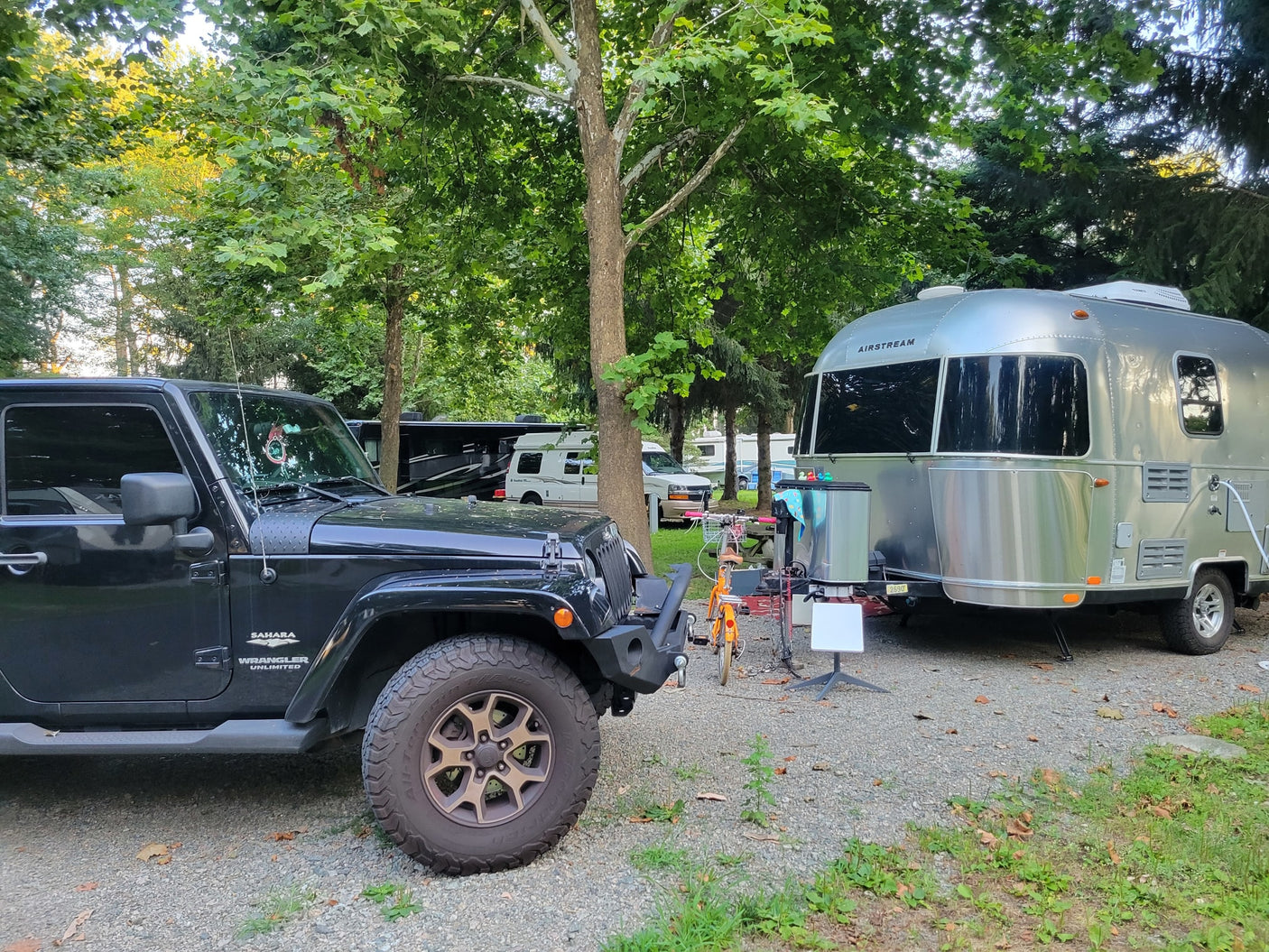 A black Jeep and a shiny silver Airstream travel trailer are parked on a gravel RV rental site under tall trees, ready for adventure.