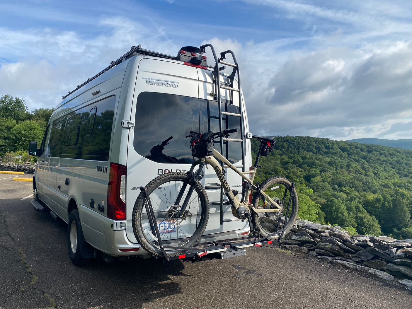 Silver Winnebago Boldt RV, fitted with a bike rack & mountain bike, parked overlooking a valley. Consider for your rv rental trip.