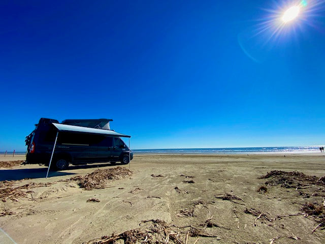 parked on a wide sandy beach by the ocean.|A dark camper van with a pop-top and awning