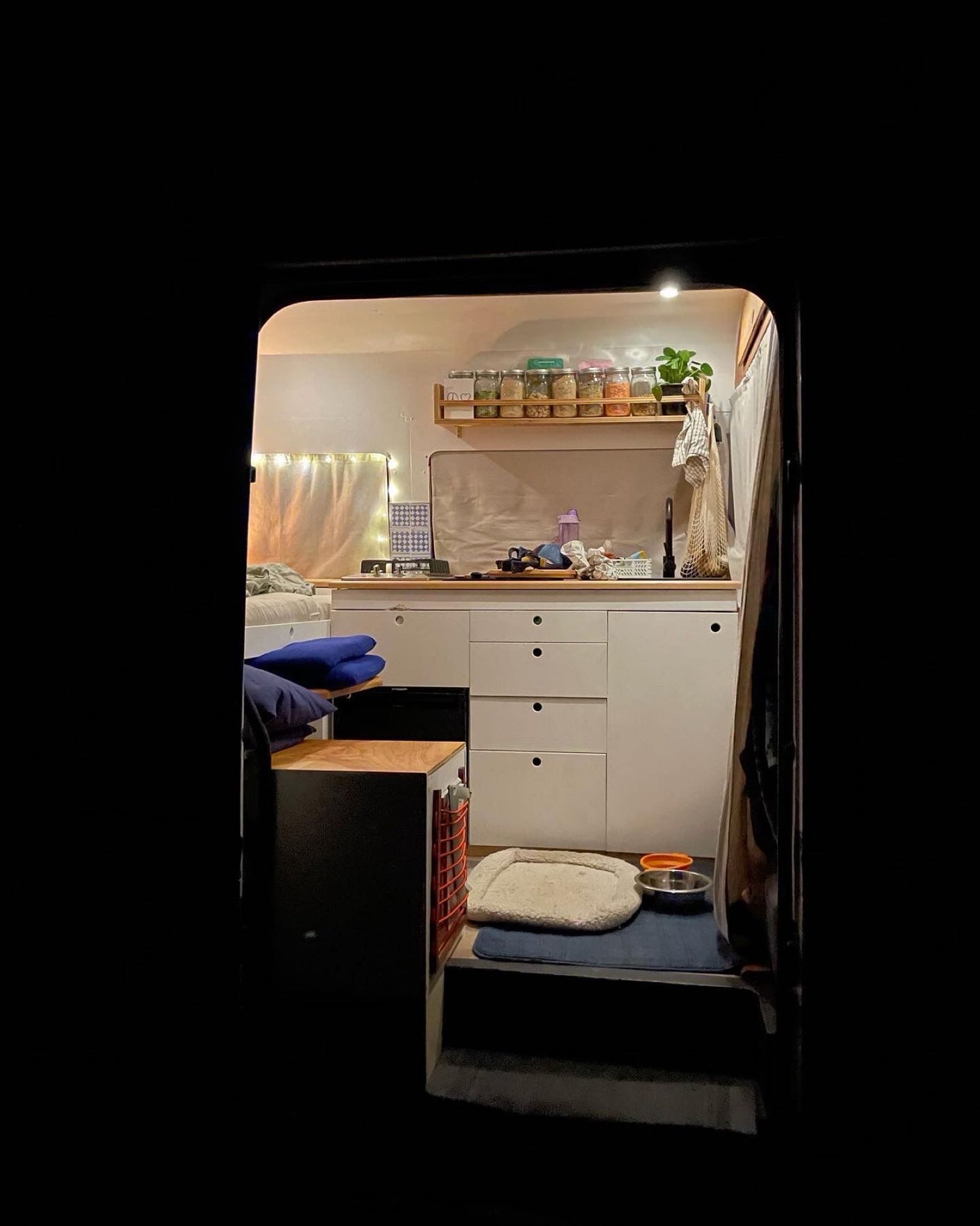 Step into this cozy RV rental interior! Features a compact kitchenette, sleeping area, and pet zone, all warmly lit at night.
