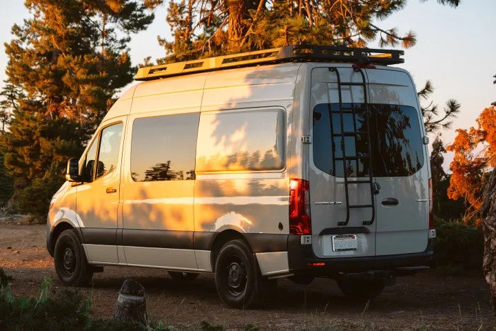 Light gray custom Sprinter van RV rental with roof rack & ladder, bathed in golden light in a forest setting.