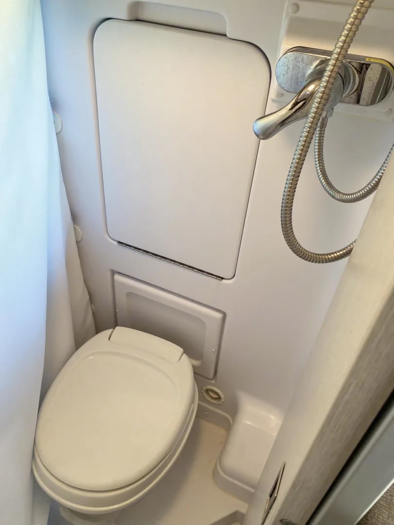 and kitchen. Ideal for comfortable road trip adventures.|This RV rental features a comfortable lounge area with cup holders and serene nature views from its large windows.|An RV rental's compact wet bath showing a white toilet