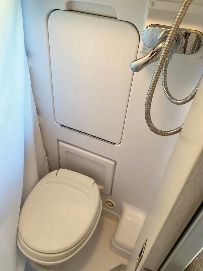 and kitchen. Ideal for comfortable road trip adventures.|This RV rental features a comfortable lounge area with cup holders and serene nature views from its large windows.|An RV rental's compact wet bath showing a white toilet