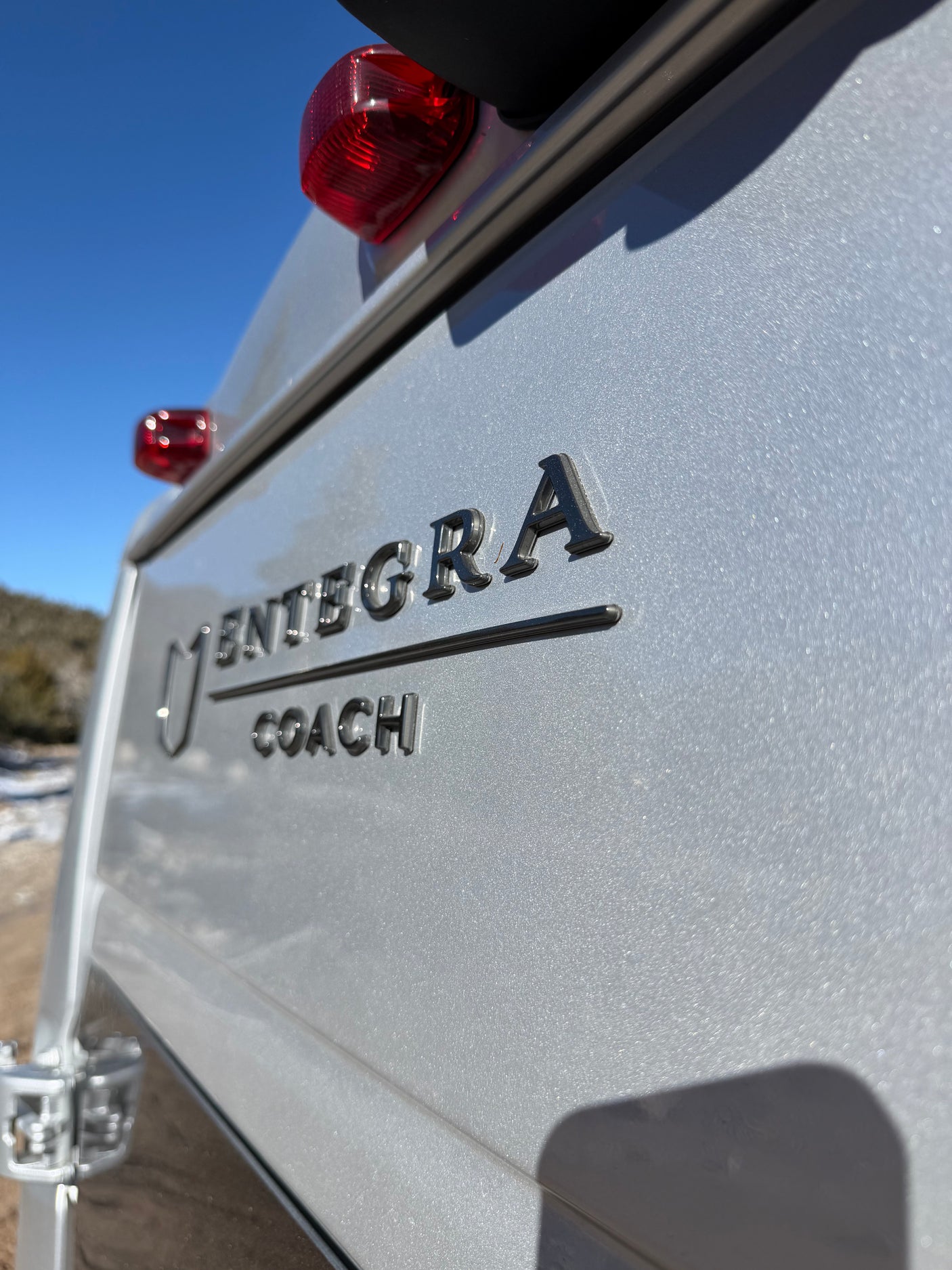 Close-up of a silver Entegra Coach RV's rear, featuring raised brand letters and red taillights. Great for your next rv rental journey.