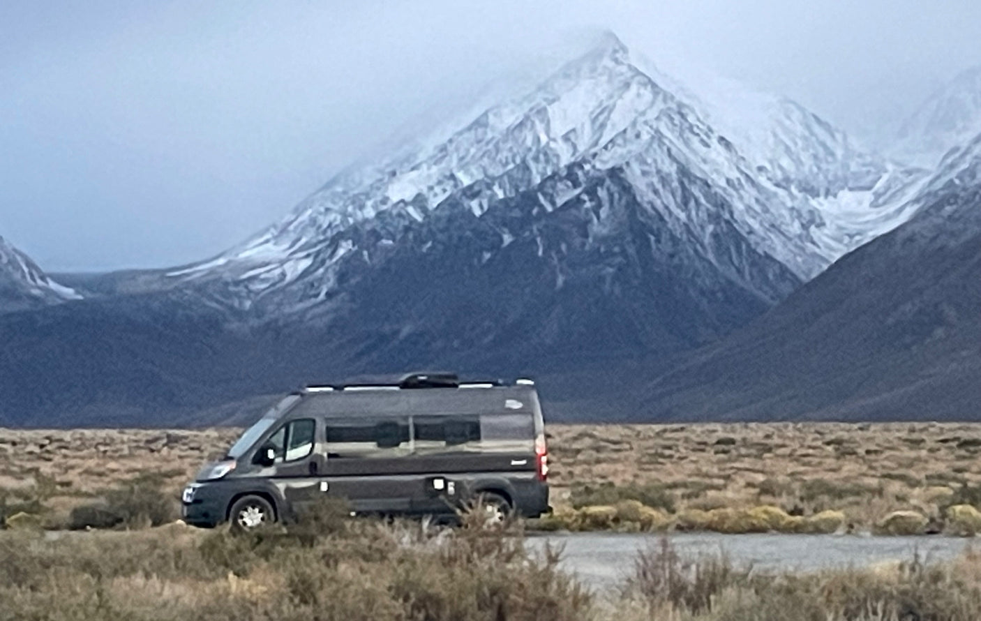 Gray RV rental parked on a vast plain with dramatic snow-capped mountains. Ideal for scenic road trips and wilderness adventures.