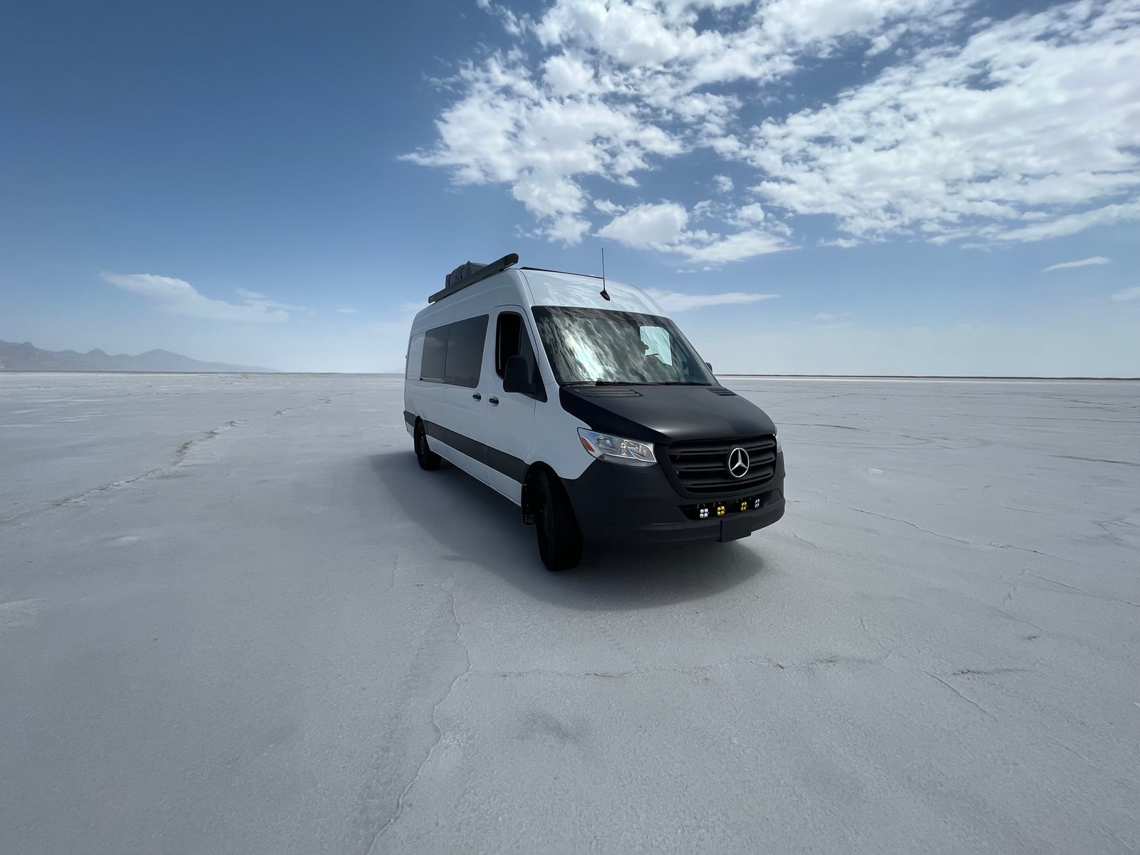 A white and black RV rental van parked on a vast salt flat, with distant mountains and a blue sky overhead.