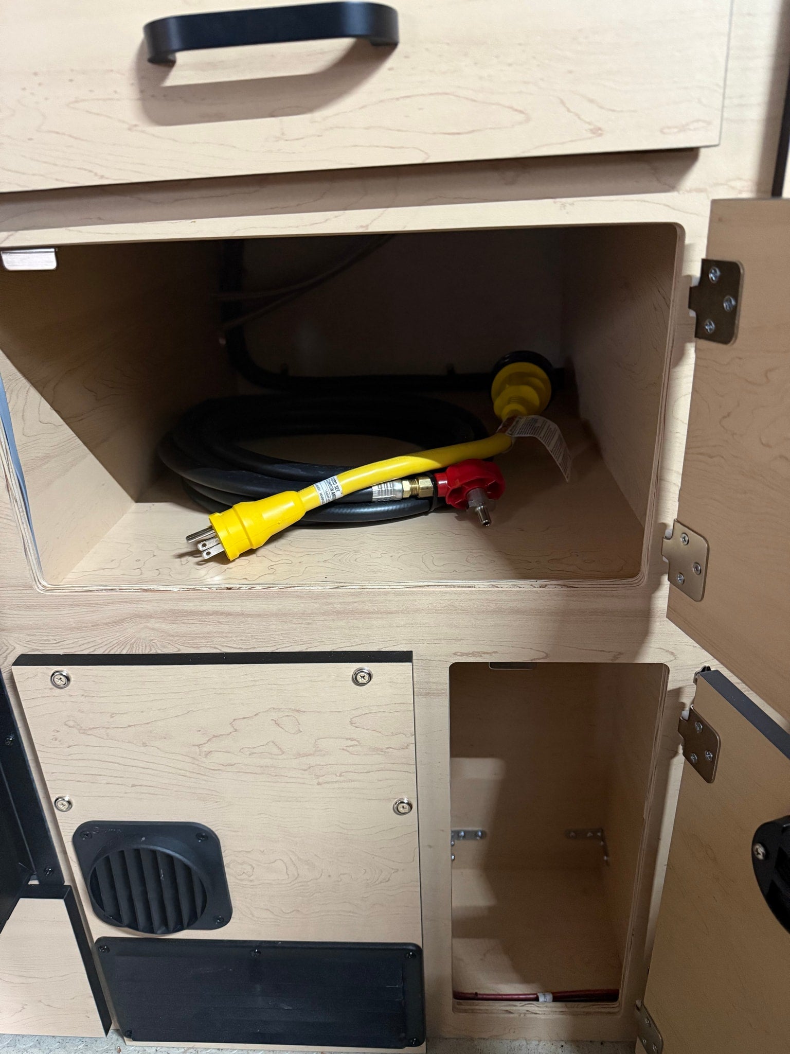 RV rental storage: an open cabinet reveals a coiled black electrical cord, a yellow adapter, and a red gas line connector.