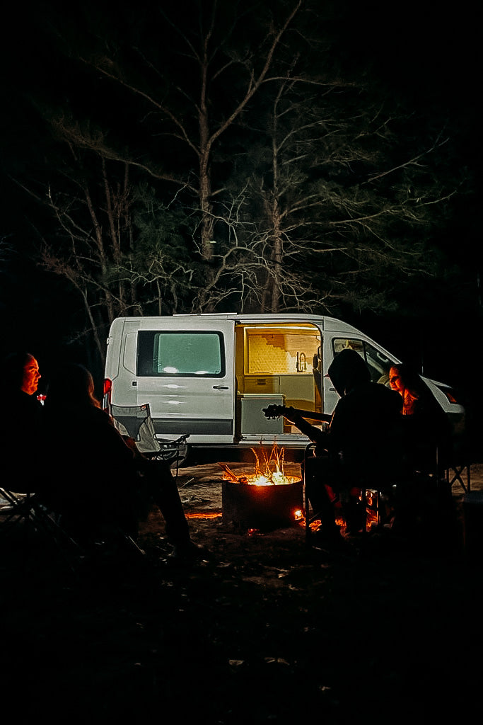 Night scene: Friends gathered around a bright campfire with a guitar, next to a cozy rv rental camper van.