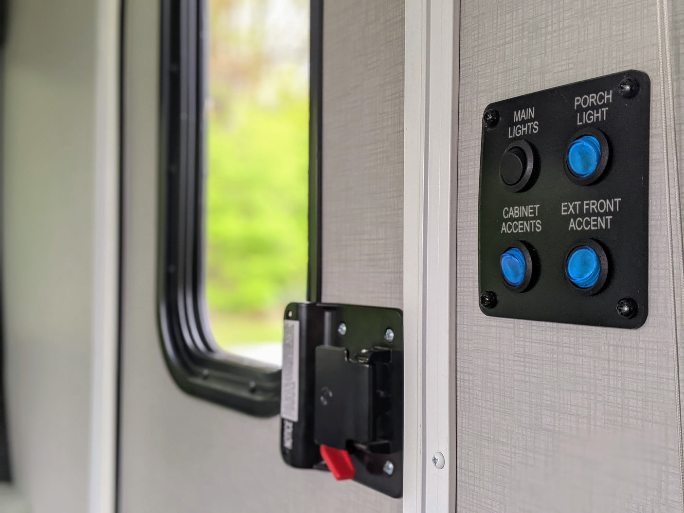 RV rental interior: Light control panel with blue buttons for porch, cabinet, and exterior accent lights, plus a black main light switch, beside a window.