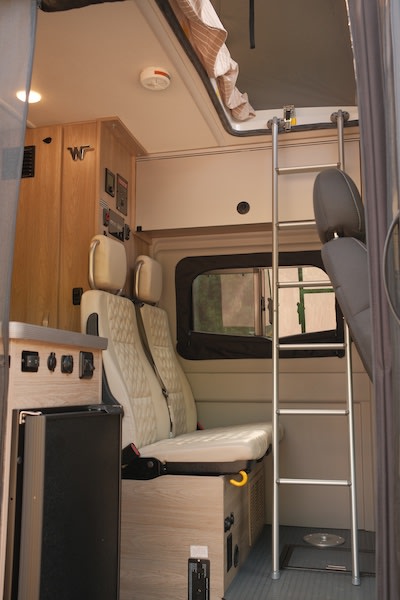 Inside a compact RV rental, showing beige quilted seats, a fridge, control panels, and a ladder to the pop-top sleeper.