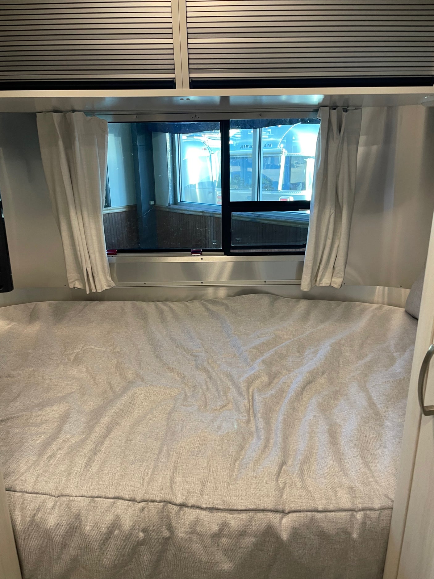 RV rental interior: cozy bed with open curtains, revealing Airstream trailers outside a large window. Overhead blinds are closed.