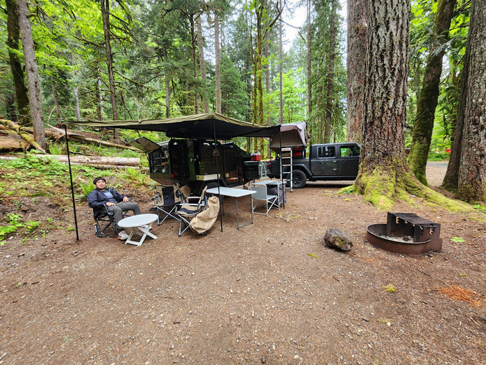 An RV rental overlanding adventure at a forest campsite. A man relaxes beside a rugged trailer & Jeep with a rooftop tent.