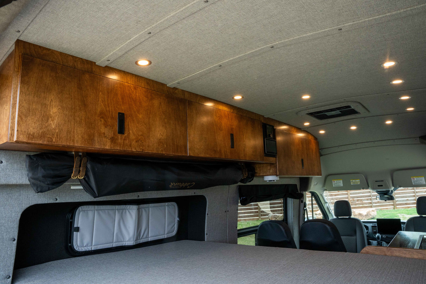 Custom wood cabinetry and bright recessed lighting illuminate the comfortable bed area of this RV rental camper van.