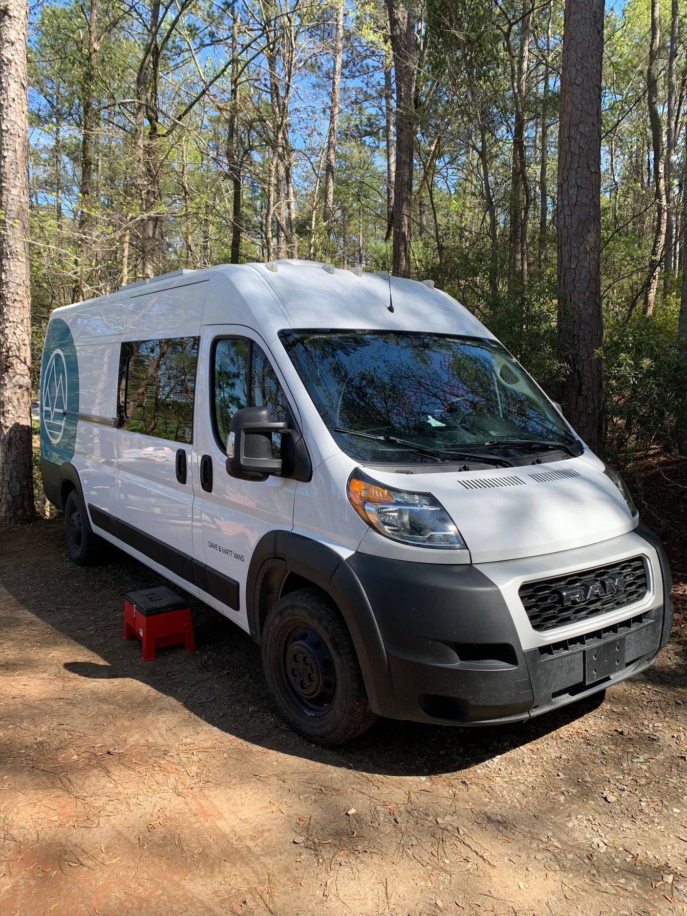 A white "Dave & Matt Vans" camper van (RV rental) is parked on a dirt path in a sunny forest, with a red step stool beside it.