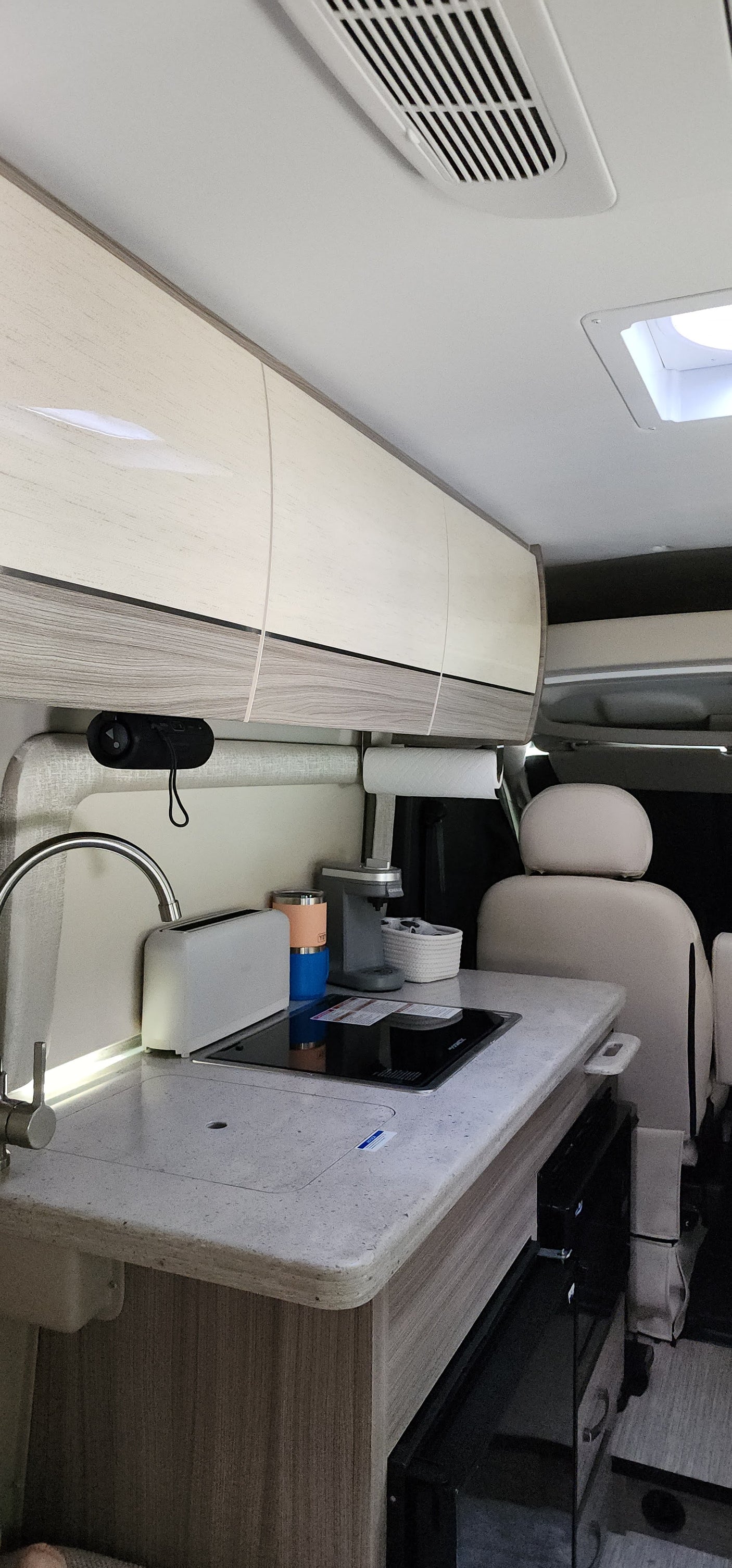Inside this RV rental, find a compact kitchen: sink, induction cooktop, coffee maker, & overhead cabinets. Ready for adventure.