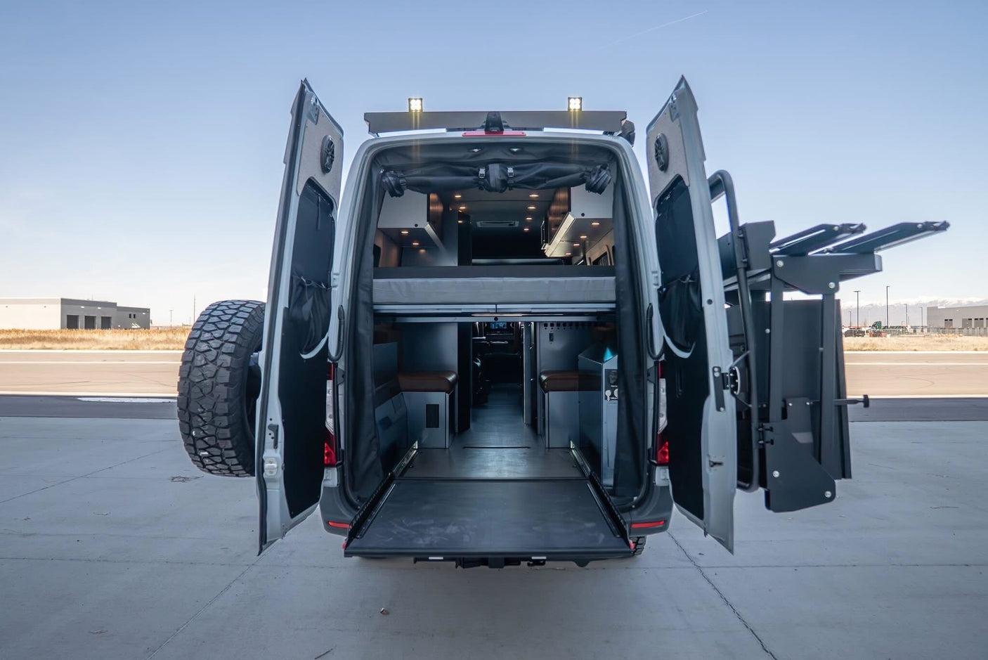 Adventure awaits! This **rv rental** camper van shows its custom interior with a bed, seating, pull-out deck, and rear-mounted gear/tire racks.