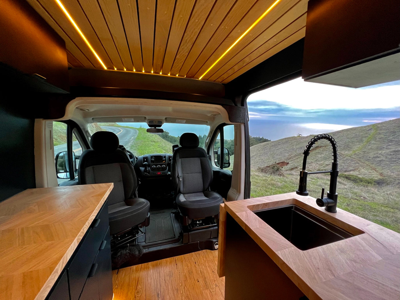 Modern RV rental interior with illuminated wood ceiling, light countertops, sleek black sink, and scenic views.