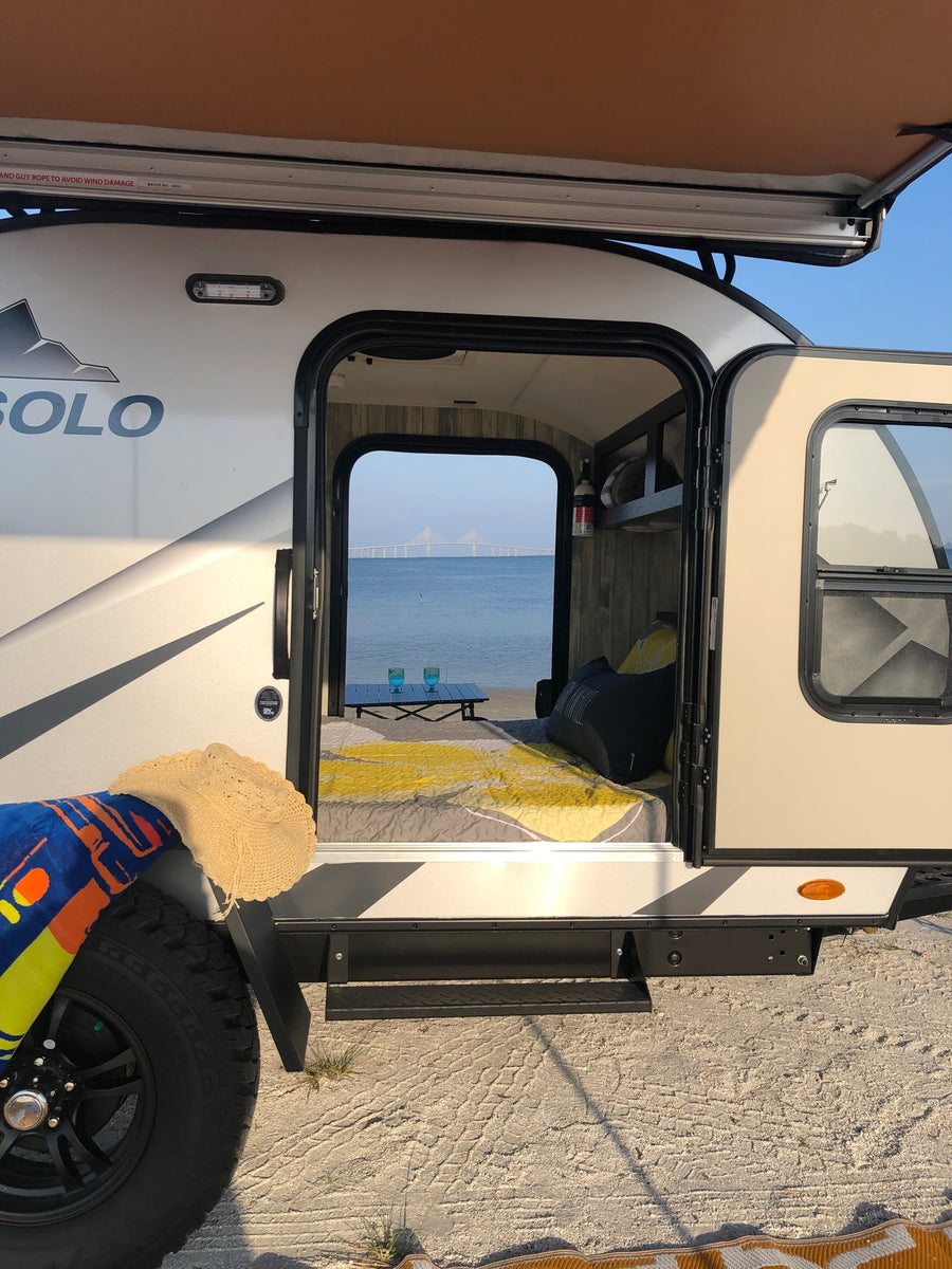 RV rental: Camper on sandy shore, open door reveals cozy bed, table, and stunning bridge view over water.