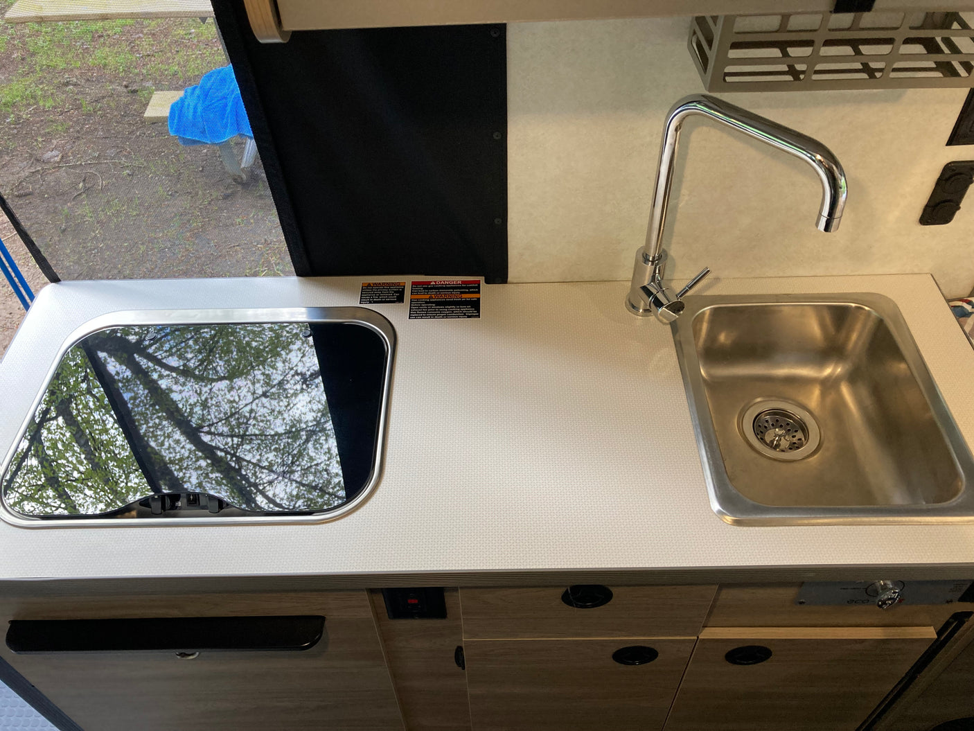 Compact rv rental kitchen with stainless steel sink, chrome faucet, and covered cooktop reflecting trees.