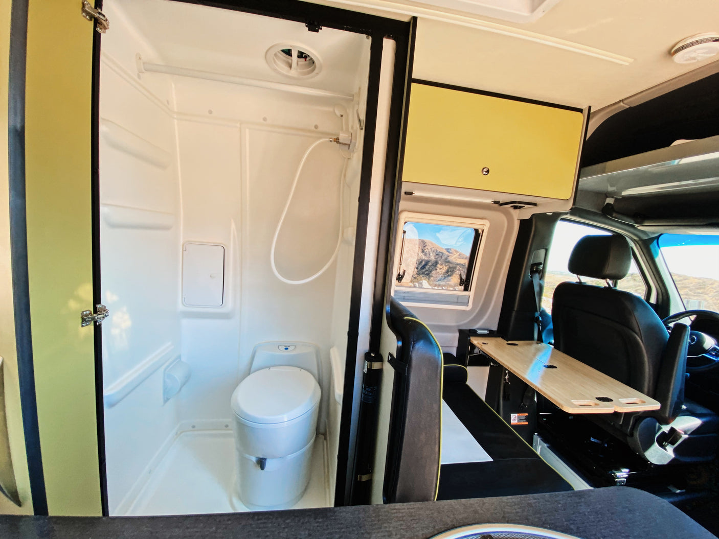 RV rental interior: compact wet bath (toilet, shower) and dining area with table, overlooking a mountain landscape.