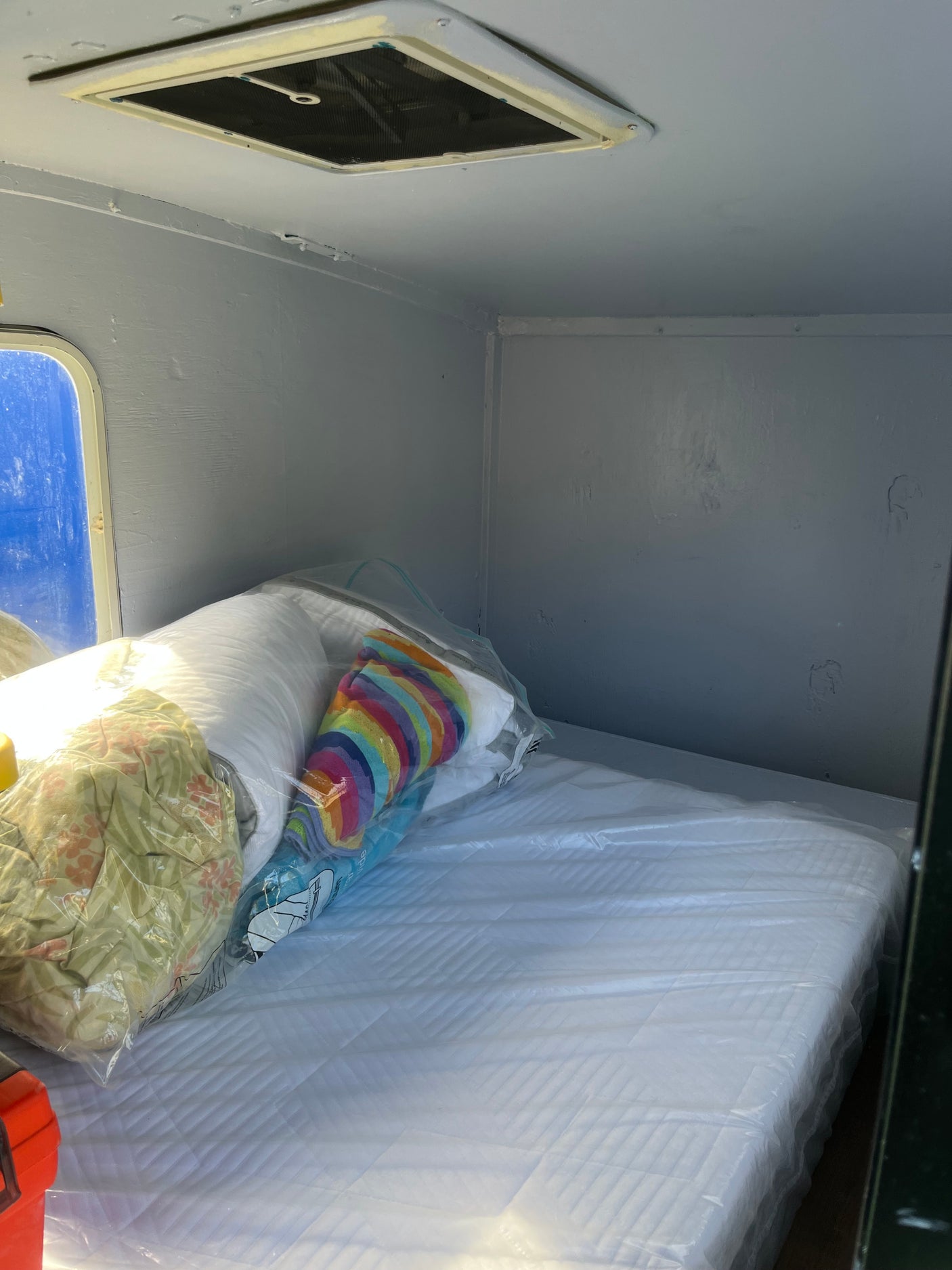 Clean RV rental sleeping area: new mattress in plastic, pillows, window, and ceiling vent in light interior.