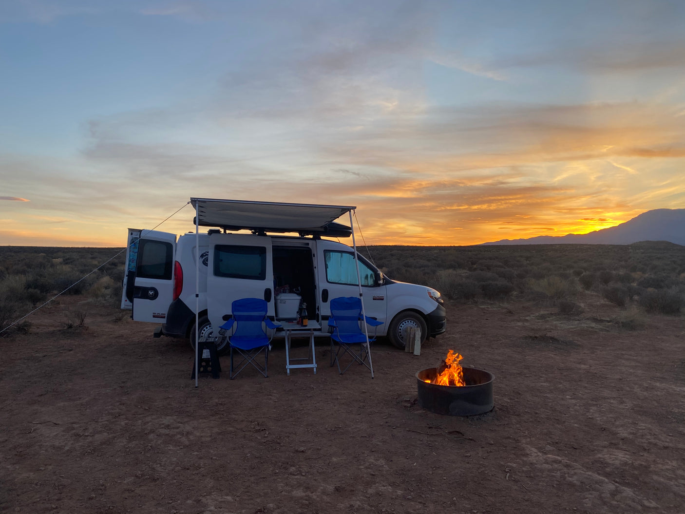 A white RV rental van with an extended awning, chairs, and campfire set against a stunning desert sunset.