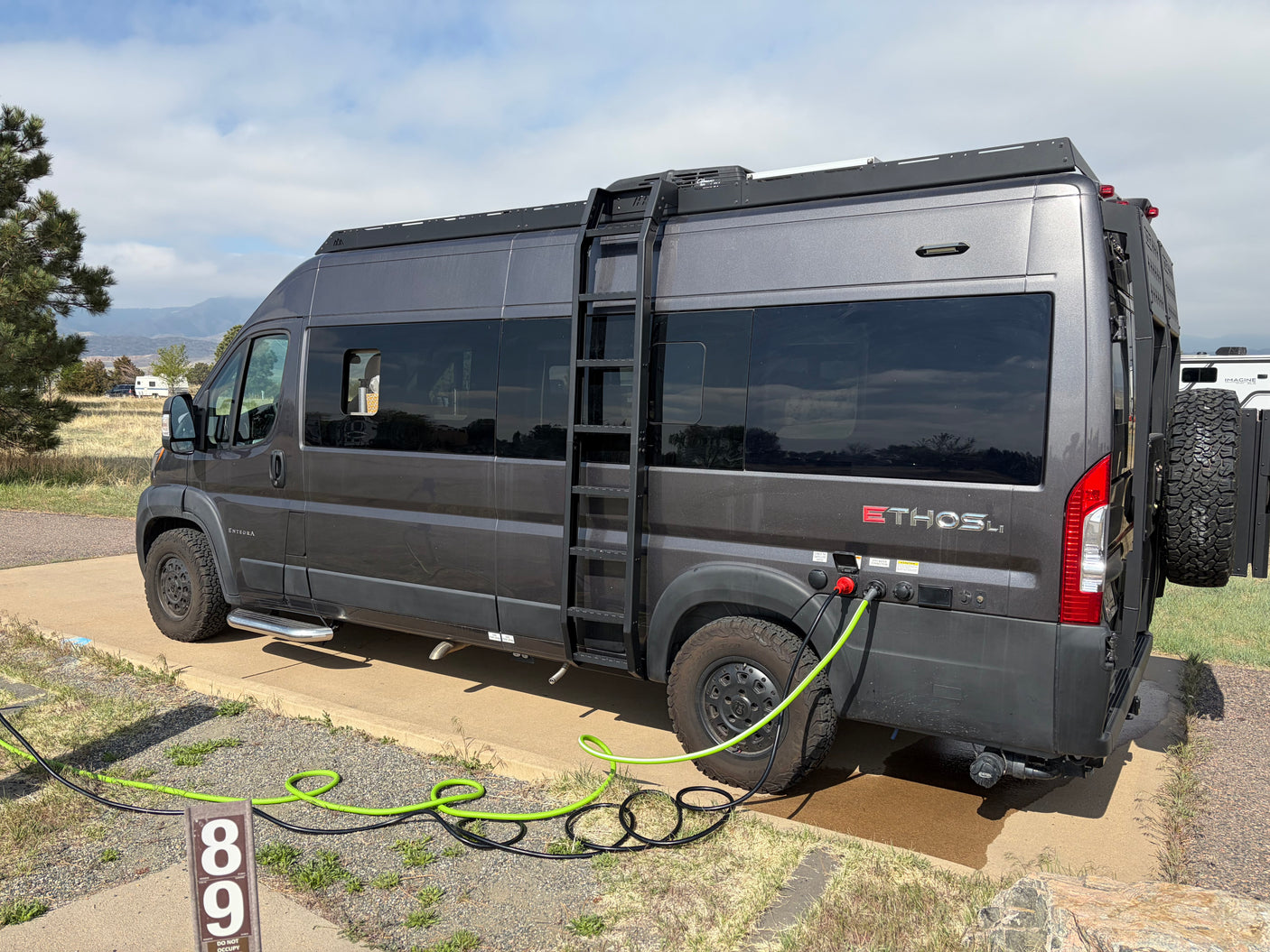 Dark grey rv rental camper van at a scenic campsite, hooked up with power and water, mountains in the distance.