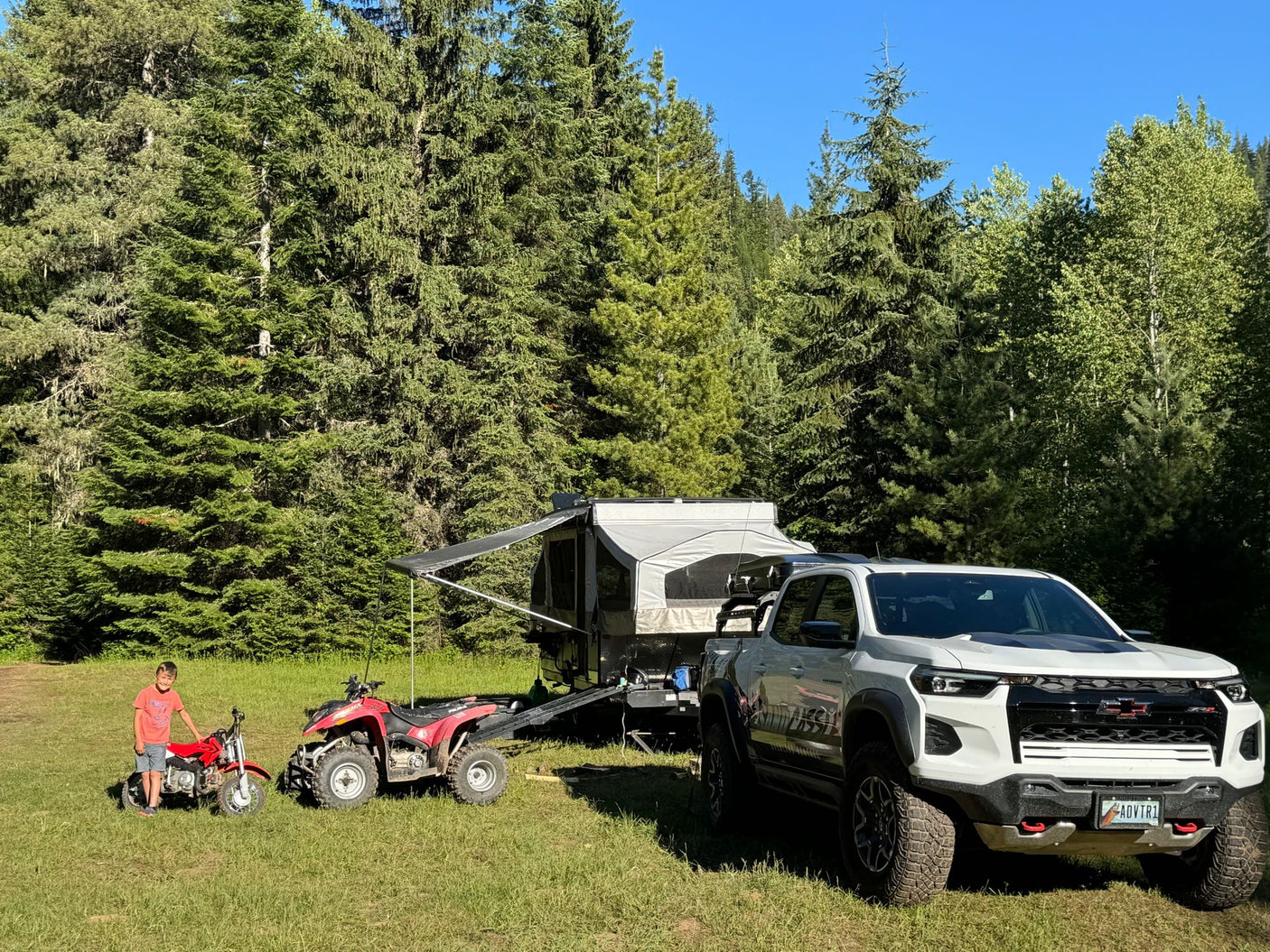 A boy with a dirt bike & ATV next to a white truck and pop-up camper. Ideal for an RV rental camping trip in the forest.