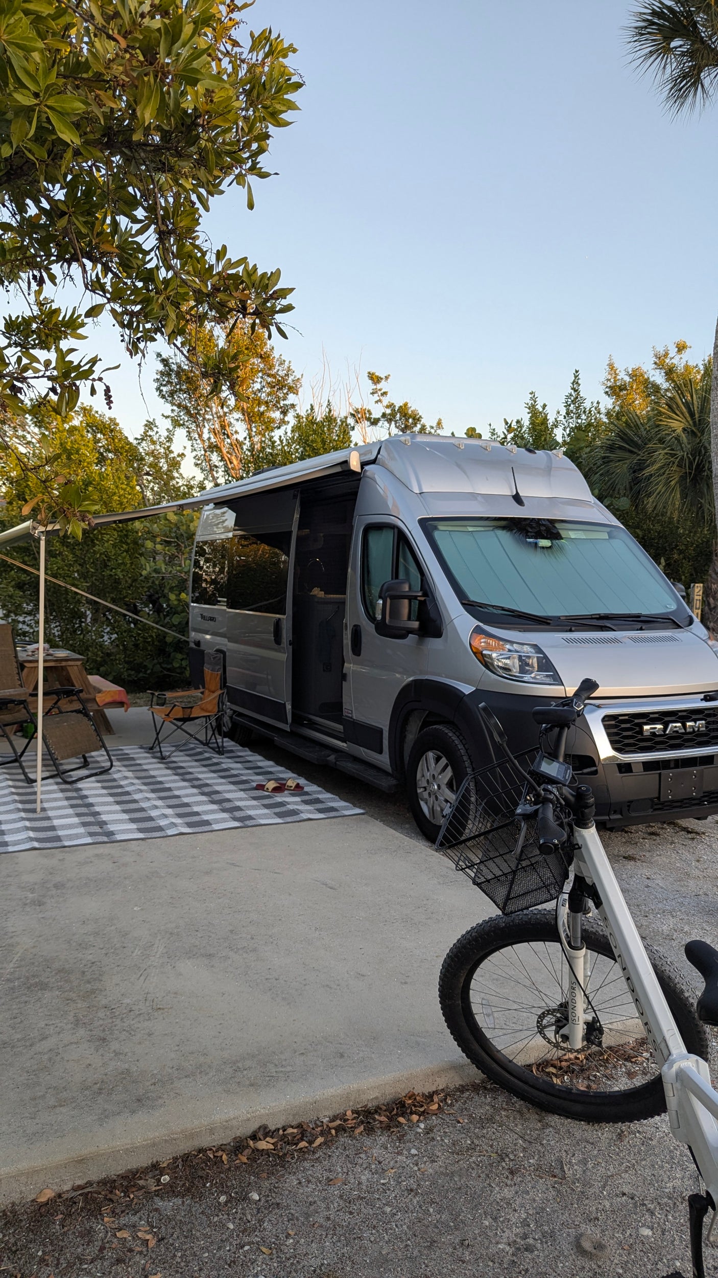 Silver camper van RV rental at campsite with extended awning, outdoor seating, and bike.