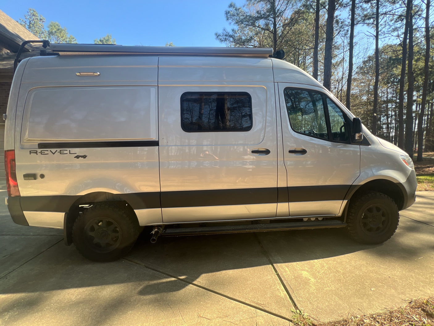 with an extended awning and trees nearby.|A silver Winnebago Revel 4x4 adventure camper van