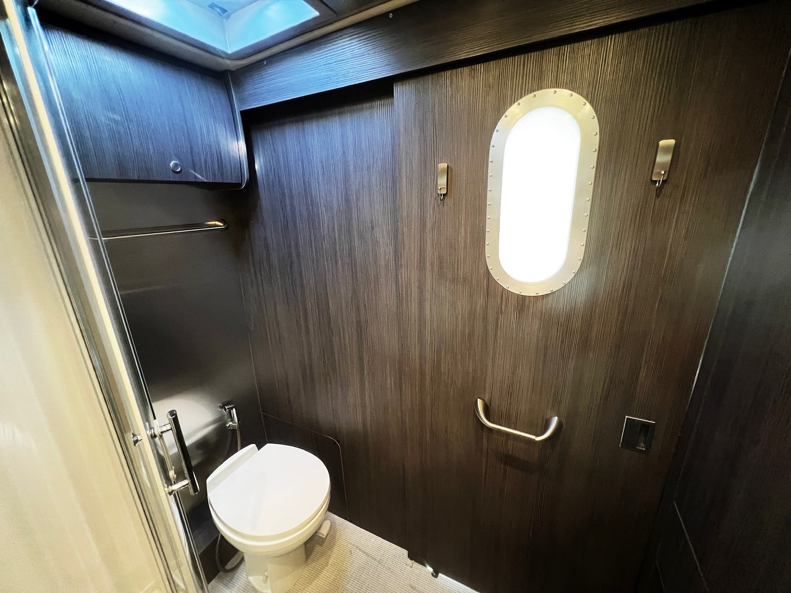 An RV rental bathroom with a toilet, shower, dark wood paneling, porthole-style light, hooks, and a grab bar.