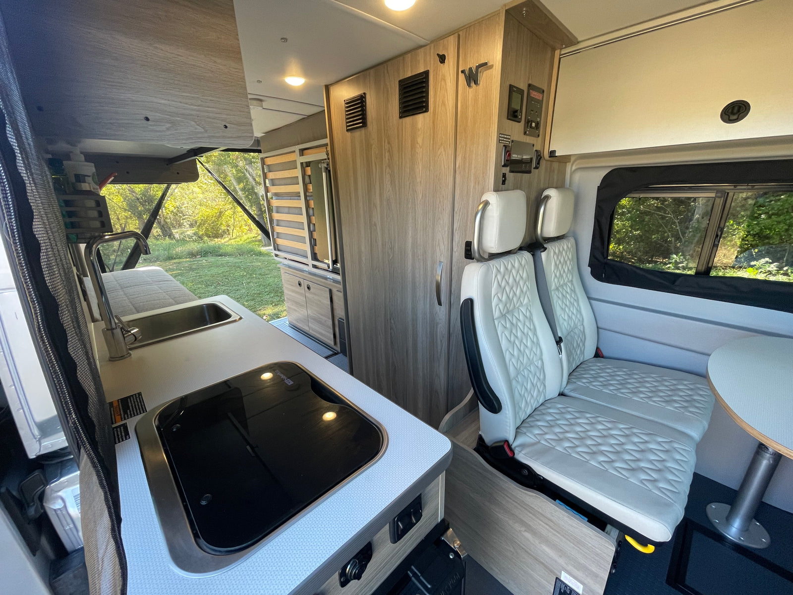 Inside a modern RV rental: kitchenette, dinette with sleek white seats, and versatile sleeping area in a bright camper van.