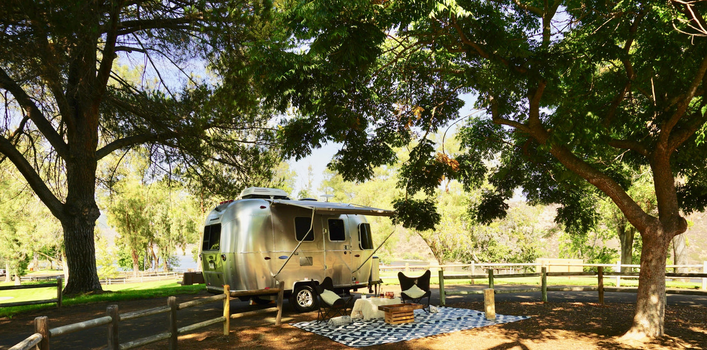 An Airstream RV rental is parked under trees, with an inviting outdoor lounge set up on a patterned rug by the water. — photo 2