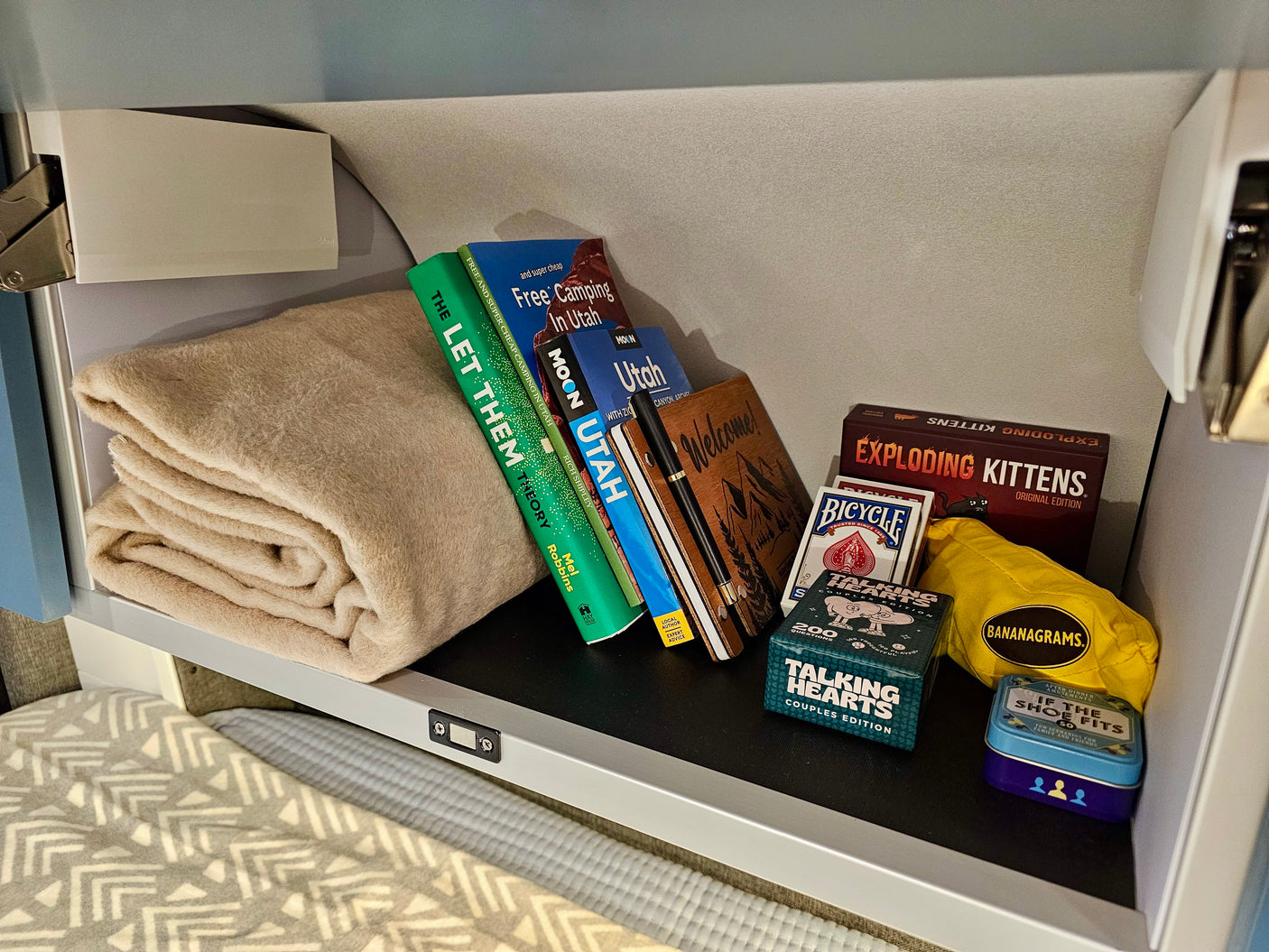 RV rental shelf holds a cozy blanket, Utah travel guides, and fun games like Exploding Kittens and Bananagrams. Adventure awaits!