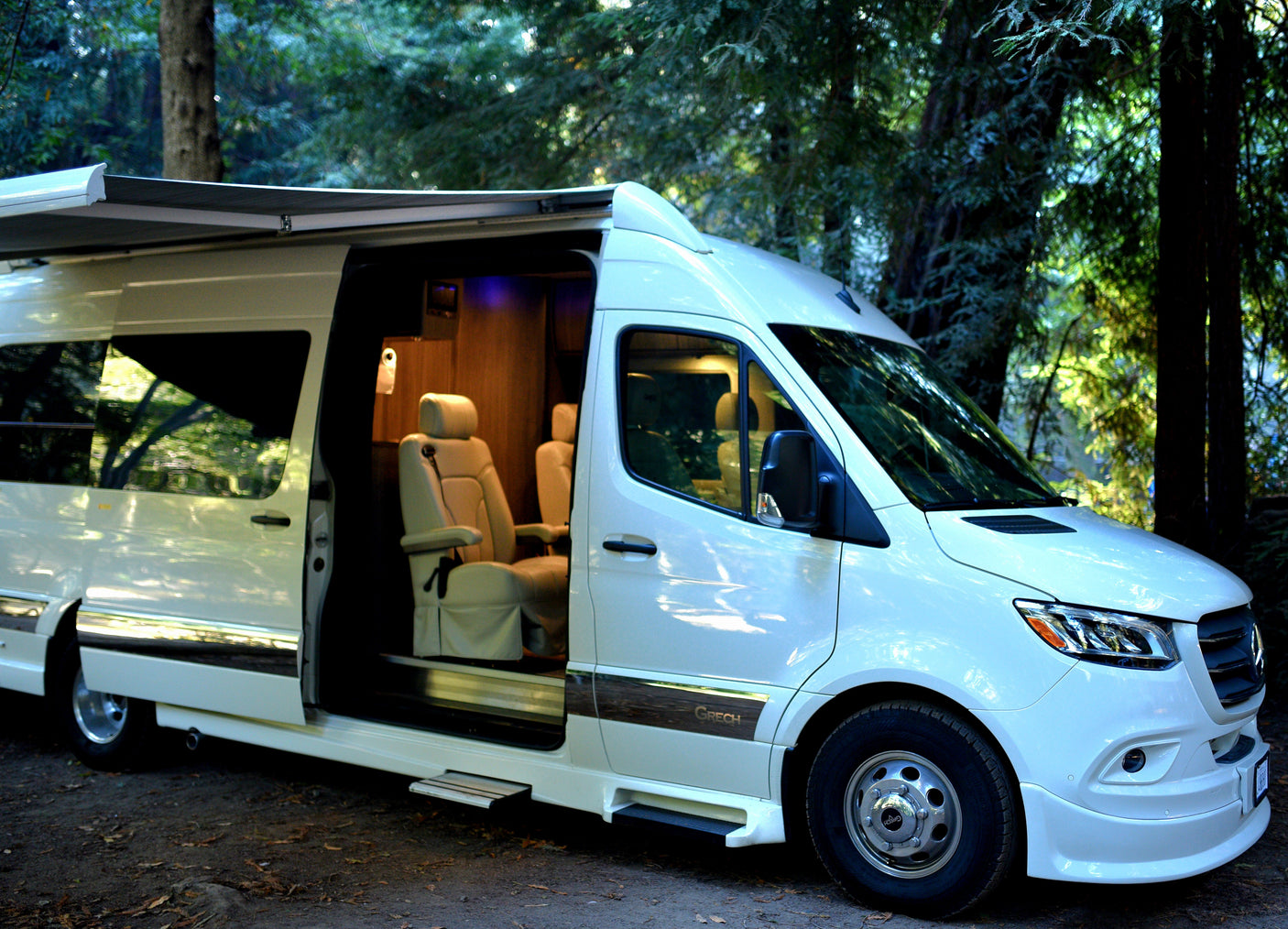 White luxury camper van RV rental with an extended awning, open door showing plush interior, parked in a shaded forest.