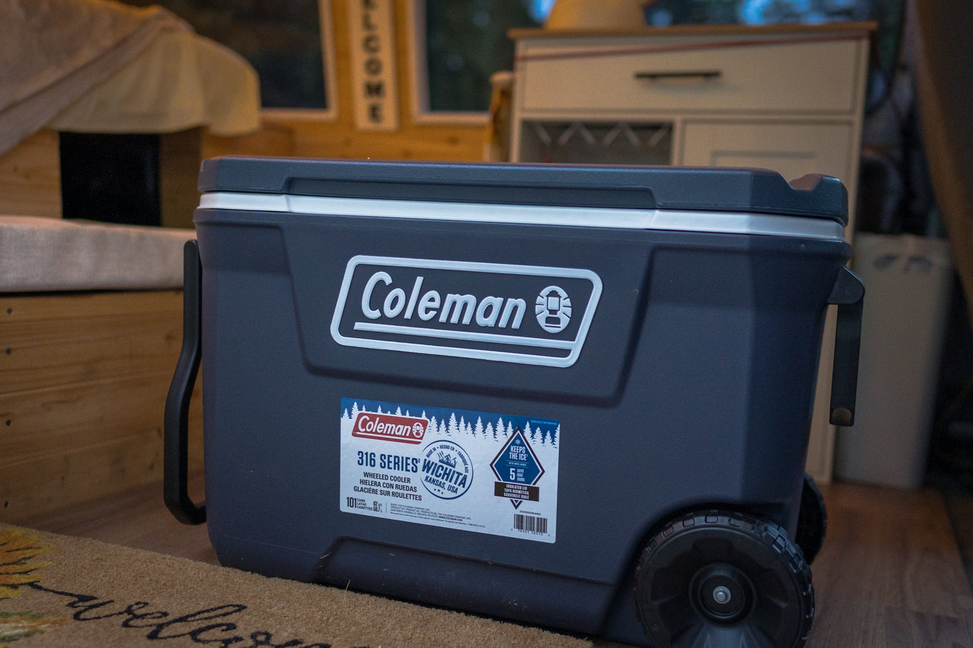 This dark grey Coleman 316 Series wheeled cooler sits on a welcome mat, perfect for your RV rental, keeping ice for 5 days.
