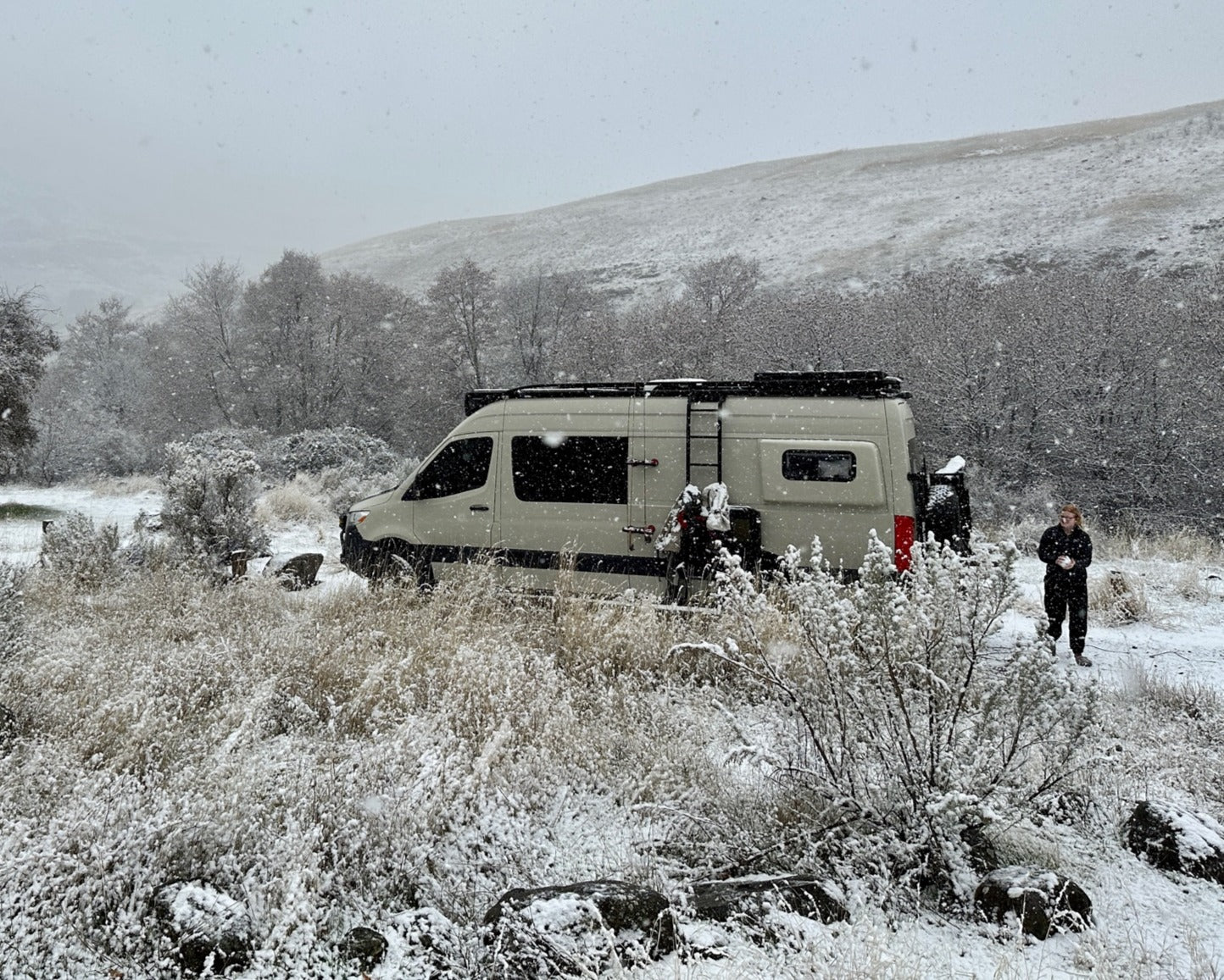 Beige campervan RV rental parked in a snowy winter landscape with a person standing nearby as snow falls lightly.