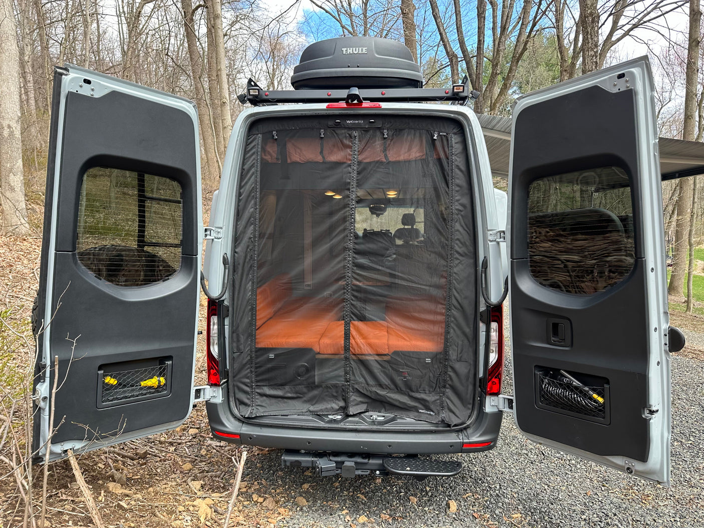 Rear of an RV rental van with doors open, revealing a bug screen and orange cushions inside, plus a Thule roof box.