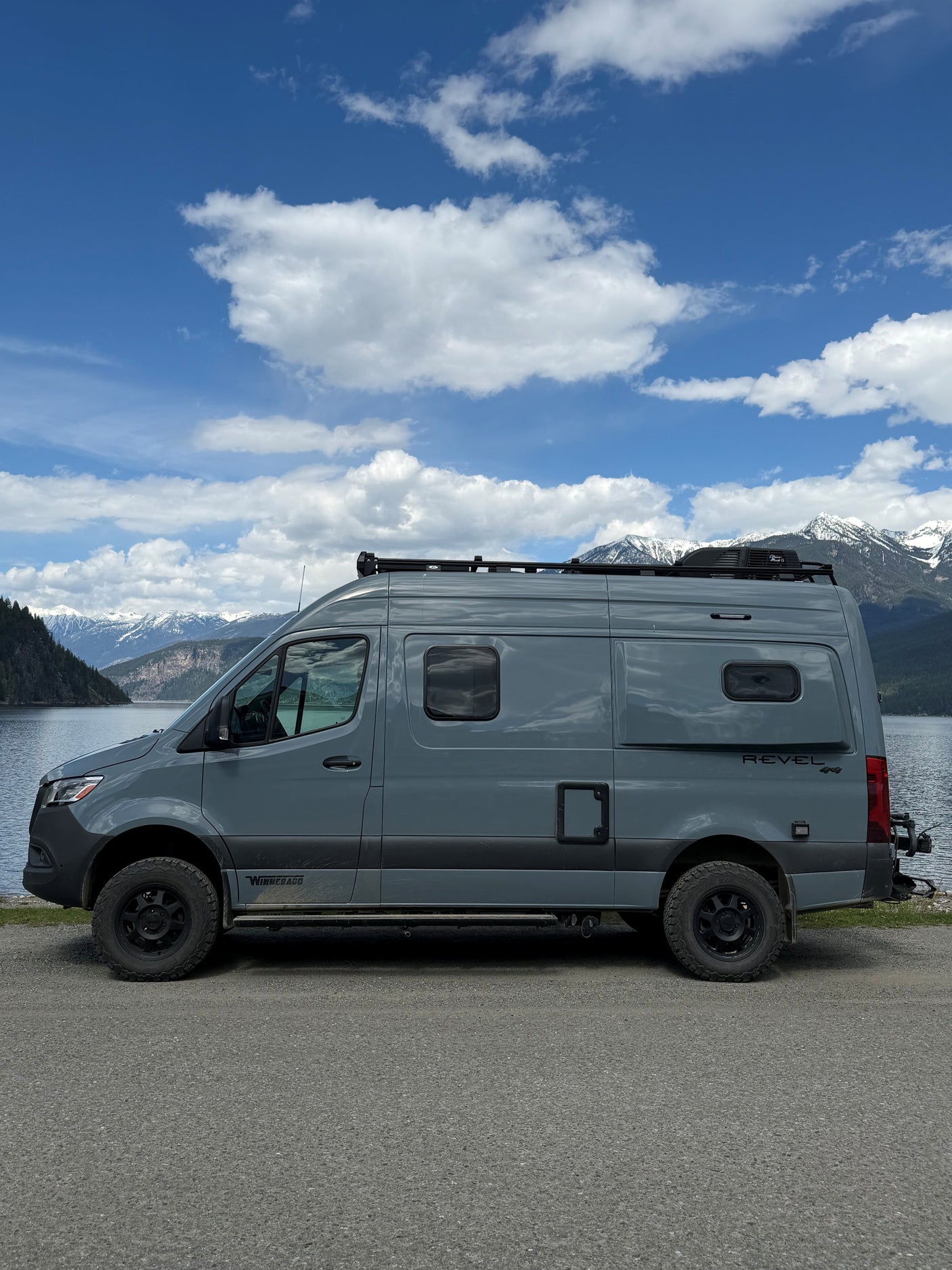 This grey Winnebago Revel 4x4 RV rental is parked by a stunning lake with snow-capped mountains and a blue sky.