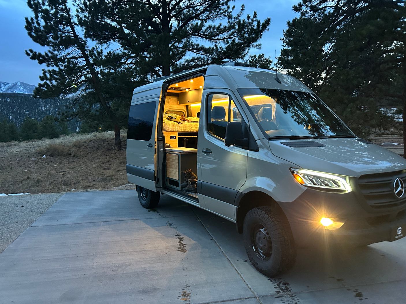 A modern **RV rental** camper van with an illuminated interior is parked at twilight, surrounded by trees and mountains. Perfect for adventure!